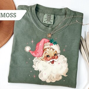 May include: A moss green t-shirt featuring a vintage Santa Claus graphic with a pink hat and white beard. The shirt has a crew neck and short sleeves. A gold necklace with a pendant is draped across the shirt.