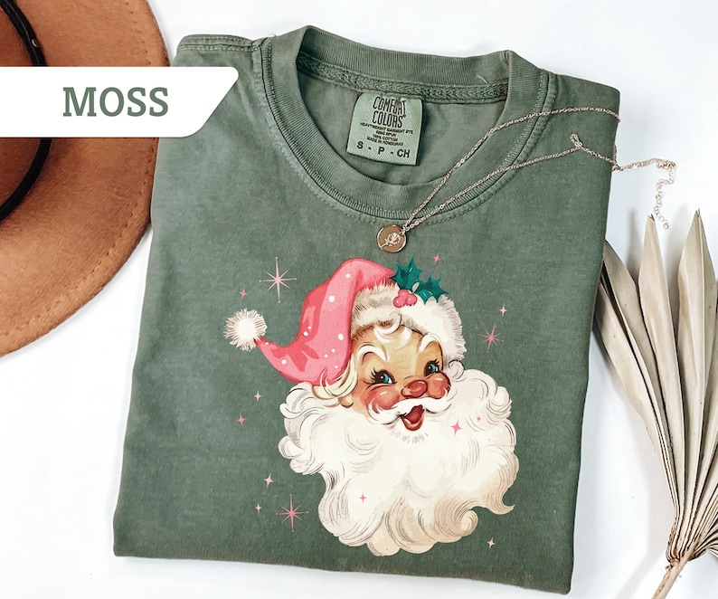 May include: A moss green t-shirt featuring a vintage Santa Claus graphic with a pink hat and white beard. The shirt has a crew neck and short sleeves. A gold necklace with a pendant is draped across the shirt.