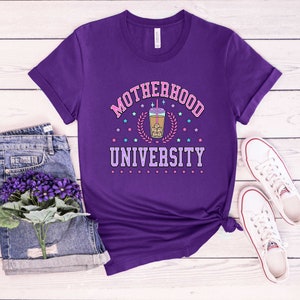 May include: Purple t-shirt with the text "Motherhood University" in pink and white letters. A cartoon image of a boba tea drink is in the center of the text.