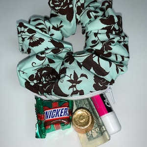 Pode incluir: Um scrunchie floral azul-turquesa e marrom com um bolso com zíper. O bolso contém uma barra de chocolate Snickers, uma moeda de chocolate dourada, uma nota de um dólar enrolada e um bálsamo labial rosa e branco.