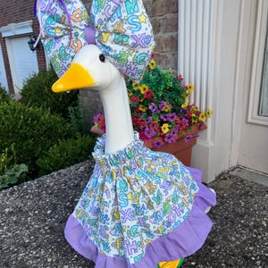 May include: A white plastic goose statue wearing a fabric outfit featuring a colorful alphabet print. The ensemble includes a ruffled skirt with a solid purple hem and a matching oversized bow headpiece adorned with a purple ribbon tie.