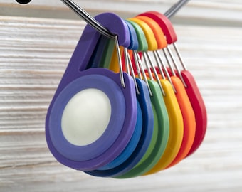 May include: A set of colorful, plastic carabiner clips with a white, round AirTag holder. The clips are arranged in a rainbow pattern, with the purple clip at the top and the red clip at the bottom. The text (AIR TAG) CARABINER SPECTRUM SERIES is printed below the clips.