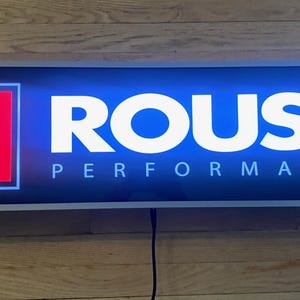 May include: Rectangular illuminated sign with the text "ROUSH PERFORMANCE" in white letters on a blue background. The left side features a red "R" logo within a white-bordered square. The sign is mounted on a wooden surface.