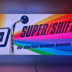 May include: A rectangular illuminated sign featuring a Hurst Super/Shifter 2 graphic. The design includes a rainbow background, a gear shifter illustration, and the text New!, HURST, SUPER/SHIFTER 2, and the shortest distance between 4 gears.
