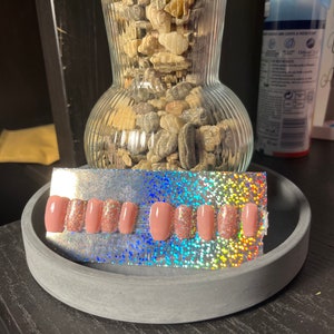 May include: A set of press-on nails in a neutral pink colour, with some nails featuring a glitter accent. The nails are displayed on a silver holographic background, presented in a grey, circular tray.