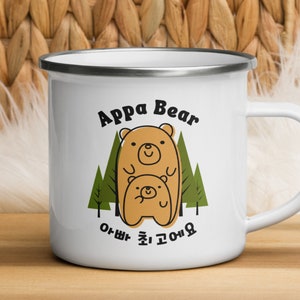 May include: White enamel mug with a silver rim. The mug features a cartoon illustration of two bears in a forest setting. The text "Appa Bear" is written above the bears and the text "아빠 춤 고마워" is written below the bears.
