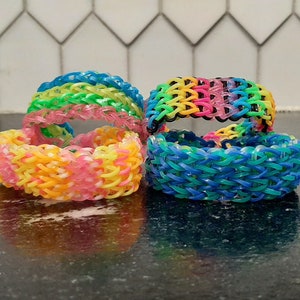 May include: Four colorful rubber band bracelets stacked on a black surface. The bracelets are made with rainbow, pink, blue, and green rubber bands.