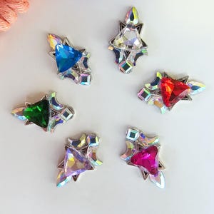 May include: Six colorful gemstone embellishments with silver-tone metal settings. Each gem features a different colored triangular center stone: blue, green, red, pink, and clear. The gems are arranged on a white surface.