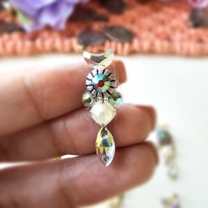 May include: A close-up of a decorative earring with iridescent rhinestones and a pearl-like bead. The earring has a crescent-shaped top, a central cluster of colorful stones, and a teardrop-shaped bottom. The design is intricate and eye-catching.