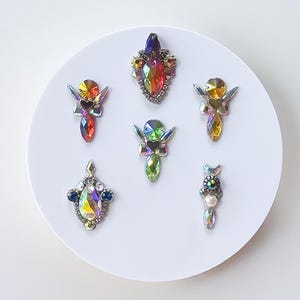 May include: A collection of iridescent rhinestone embellishments in various shapes and sizes. The rhinestones feature a range of colors, including red, orange, yellow, green, and blue. They are arranged on a white circular surface, creating a sparkling and decorative display.