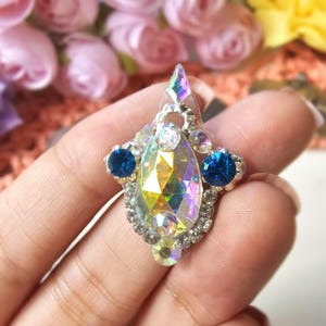 May include: A decorative jewel with a silver setting, featuring a large, iridescent, teardrop-shaped crystal as the centerpiece. Two smaller, round, deep blue crystals flank the main stone. The jewel is embellished with smaller clear crystals.