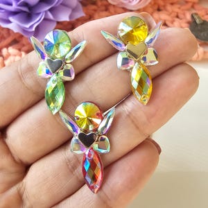 May include: Three iridescent rhinestone fairy embellishments. Each fairy has a round head, heart-shaped body, and wing-shaped accents. The color palette includes shades of green, yellow, and red, with a black heart detail. These are ideal for craft projects.