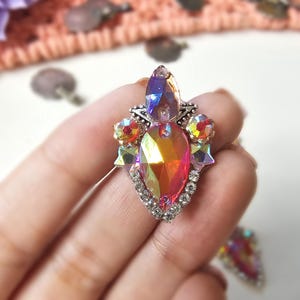 May include: A multi-faceted, iridescent rhinestone applique with a teardrop shape. The central stone is surrounded by smaller, colorful gems and a border of clear rhinestones. The design is held in a hand.