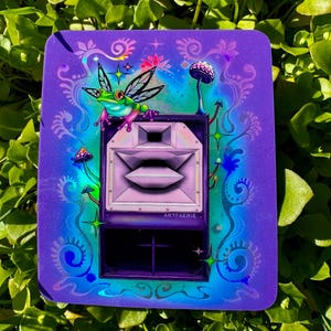 May include: A purple and blue holographic sticker with a green frog with wings sitting on top of a speaker. The speaker has a white speaker cone and the text "ARTFAERIE." is printed on the front. The sticker has a whimsical design with mushrooms and stars.
