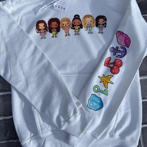 May include: White hooded sweatshirt with a colorful graphic of six cartoon women across the chest. The right sleeve features a series of colorful illustrations including a seashell, anchor, cherries, star, key, and a gem.