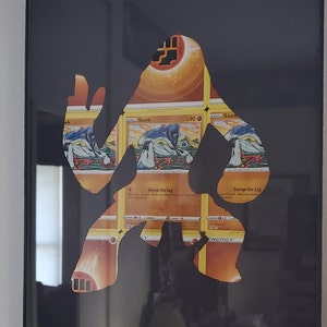 May include: A black frame with a silhouette of a Pokemon character cut out of it. The silhouette is made up of multiple Pokemon cards featuring the character Sawk. The cards are arranged in a pattern to form the silhouette. The cards feature the text "Sawk" and "Sweep the Leg".