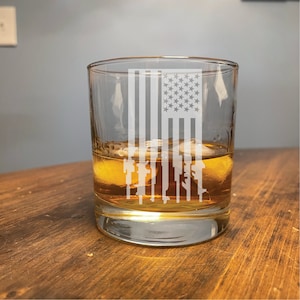 May include: A clear glass tumbler with a design of an American flag made up of guns. The glass is filled with amber liquid and ice cubes.