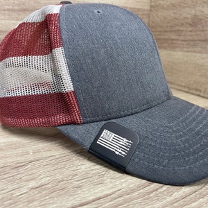 May include: A gray and red baseball cap with a mesh back. The cap has a black patch with a white American flag and gun silhouettes on the front.