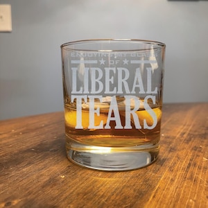 May include: A clear glass with the text "Enjoying my glass of Liberal Tears" etched on the side. The glass is filled with a brown liquid and ice cubes.