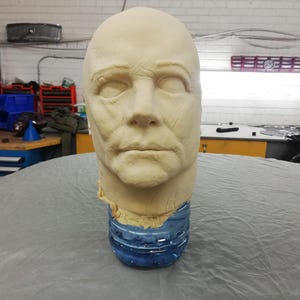 May include: A sculpted human head in a pale colour, showing detailed facial features and wrinkles. It is mounted on a blue plastic bottle. The image suggests a prop or sculpture in a workshop setting, with tools and equipment visible in the background.
