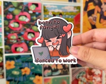 Puede incluir: Una pegatina con un personaje de estilo anime con cabello oscuro, detalles de corazón y un portátil. El texto de la pegatina dice "born to fangirl" y "forced to work". La pegatina se sostiene frente a un fondo de varias impresiones artísticas coloridas.