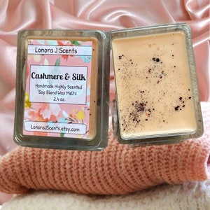 May include: A square glass container of soy wax melts with a label that reads "Lonora J Scents Cashmere & Silk Handmade Highly Scented Soy Blend Wax Melts 2.4 oz. LonoraJScents.etsy.com". The wax melts are a light peach color with black flecks.