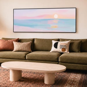 May include: A rectangular wall art piece featuring a horizontal landscape painting of a sunset over a calm ocean with soft pastel pink, blue, and white hues. The artwork is enclosed in a thin black frame and displayed above a long olive green sofa.