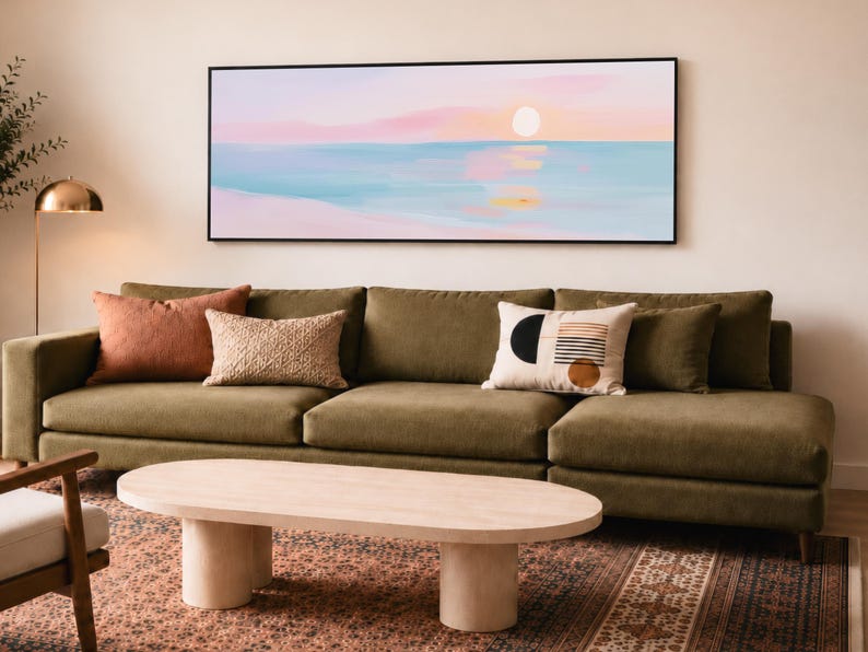 May include: A rectangular wall art piece featuring a horizontal landscape painting of a sunset over a calm ocean with soft pastel pink, blue, and white hues. The artwork is enclosed in a thin black frame and displayed above a long olive green sofa.