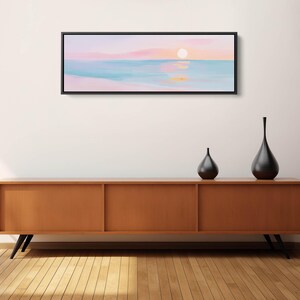 May include: A panoramic landscape painting in a black frame depicting a sunset over a calm ocean with soft pink, peach, and light blue hues. The artwork features a glowing sun reflecting on the water surface, creating a serene and horizontal composition.
