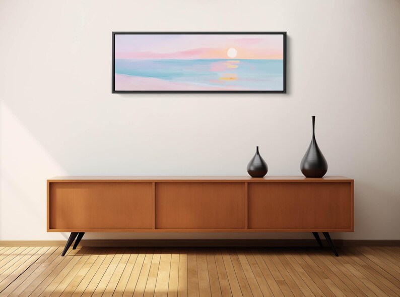 May include: A panoramic landscape painting in a black frame depicting a sunset over a calm ocean with soft pink, peach, and light blue hues. The artwork features a glowing sun reflecting on the water surface, creating a serene and horizontal composition.