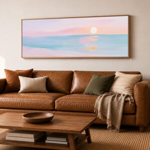 May include: A panoramic landscape painting in a light wood frame depicting a sunset over a calm ocean. The artwork features soft pastel hues of pink, peach, and light blue, with a pale sun reflecting on the water surface above a sandy beach.