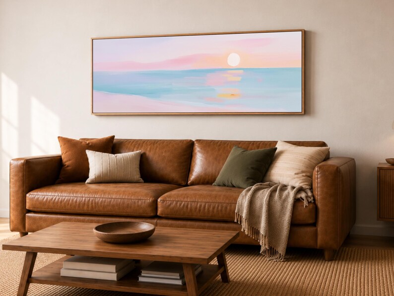May include: A panoramic landscape painting in a light wood frame depicting a sunset over a calm ocean. The artwork features soft pastel hues of pink, peach, and light blue, with a pale sun reflecting on the water surface above a sandy beach.