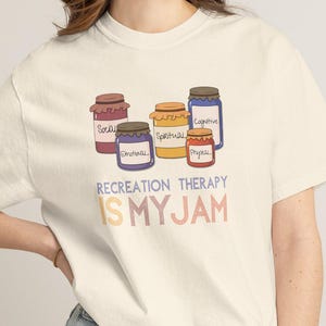 May include: A cream-colored short-sleeve t-shirt featuring a graphic of five colorful jars labeled with words including Social, Emotional, Spiritual, Physical, and Cognitive. Below the jars, the text reads RECREATION THERAPY IS MY JAM in blue and orange.