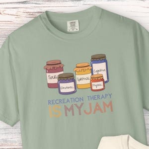 May include: A sage green t-shirt and a cream t-shirt, each featuring a graphic of five colorful jars labeled Social, Emotional, Spiritual, Cognitive, and Physical. Below the jars, the text reads RECREATION THERAPY IS MY JAM in blue and orange.
