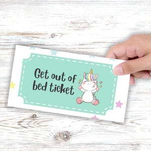 May include: A white rectangular ticket with a light teal border and the text "Get out of bed ticket". The ticket features a cartoon unicorn with a rainbow mane and horn. The background is a light wood grain.