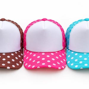 May include: Three trucker hats featuring a solid white front panel and a mesh back. Each hat has a colored brim and back with a white polka dot pattern. The hats are brown, hot pink, and bright turquoise, each with a matching button on top.
