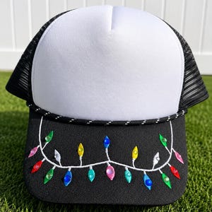 May include: A trucker hat with a white foam front panel, black mesh back, and a black brim. The brim features an embroidered white string of lights with colorful, faceted plastic bulb-shaped embellishments in red, green, blue, yellow, and pink.