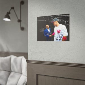 Puede incluir: Un p&oacute;ster de pared rectangular con una fotograf&iacute;a en blanco y negro de un jugador de b&eacute;isbol con camiseta de rayas estrechando la mano de una persona con traje azul. La imagen incluye dos firmas distintas en tinta azul.