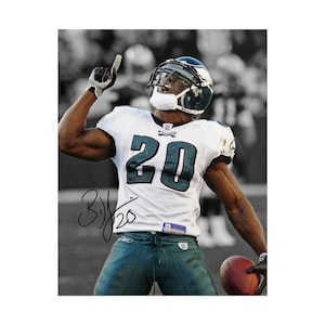 May include: A professional football player wearing a white jersey with the number 20 in dark green and black, dark green pants, and a helmet with a face mask. The player is holding a brown football and has a black ink autograph on the jersey.