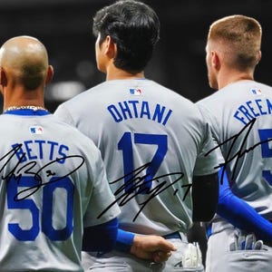 May include: A grayscale image of three baseball players from the back, wearing gray jerseys with blue accents. The jerseys display the names and numbers of Betts (50), Ohtani (17), and Freeman (5), with their signatures. The players are standing together.
