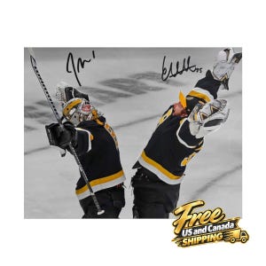 May include: A photograph of two ice hockey players in black, yellow, and white jerseys celebrating on the ice. The players are wearing goalie equipment, including masks and gloves, with their arms raised. Two distinct black ink signatures are visible.