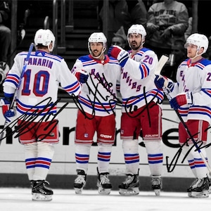 May include: Four hockey players wearing blue, white, and red jerseys with the word "Rangers" on the front. The players are all smiling and holding their hockey sticks. The players are standing on a hockey rink. The players' names are written in black marker on the jerseys.