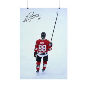 Op de afbeelding: Een verticale poster met een ijshockeyspeler van achteren gezien, gekleed in een rood shirt met de naam KANE en het nummer 88 in het wit. De speler houdt een ijshockeystick in de lucht tegen een witte ijsachtergrond, met een handtekening in zwarte inkt.