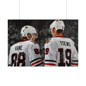 Op de afbeelding: Een foto van twee ijshockeyspelers in witte Chicago Blackhawks-shirts met zwarte en rode accenten. De speler links draagt KANE en 88 op de rug, terwijl de speler rechts TOEWS en 19 draagt. Beide shirts zijn voorzien van handtekeningen.