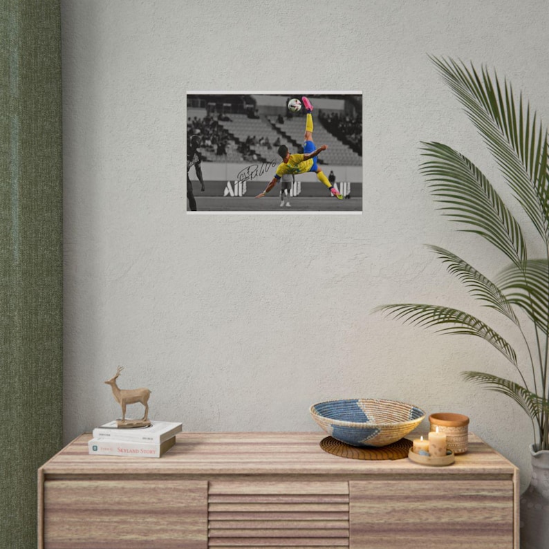 May include: A rectangular photographic print featuring a soccer player in a yellow jersey and blue shorts performing a bicycle kick. The background is a black and white stadium scene with a signature visible on the field. The print is framed in white.