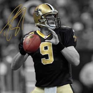May include: A football player wearing a black and gold jersey with the number 9 on the front. He is holding a red football in his left hand and is looking to the right. The background is blurred and in black and white.