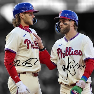 May include: Two baseball players wearing white and red Philadelphia Phillies jerseys with their signatures. The player on the left is wearing a blue baseball cap and the player on the right is wearing a blue baseball cap with a red brim.
