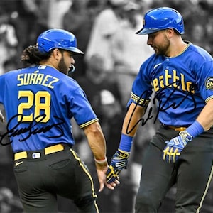 May include: Two baseball players in blue jerseys and helmets, with the word "Seattle" on one jersey. One jersey has the number 28. Both players are wearing gloves and wristbands. The background is a blurred black and white image of a crowd.