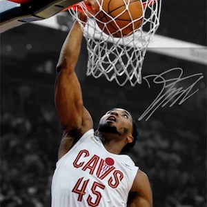 May include: A basketball player wearing a white jersey with "CAVS 45" on the front, dunking a basketball through a hoop. The player is wearing a black headband and has a determined expression on his face.