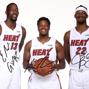 May include: Three basketball players wearing white jerseys with the word "HEAT" and their jersey numbers on the front. The players are standing in a row, with the player in the middle holding a basketball. The players' names are signed on their jerseys.